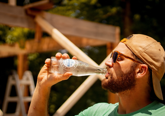 Picture of a male in a cap standing in profile drinking a glass bottle of hard seltzer on a summers day, with the seltzer bottle perspiring and glistening in the sunshine