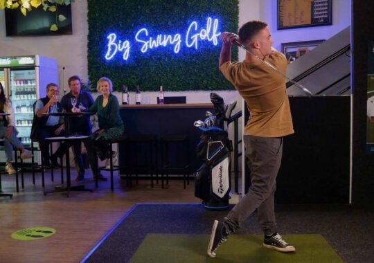 Big Swing Golf - golfer swinging a golf club with neon sign behind saying "Big Swing Golf" as friends watch on